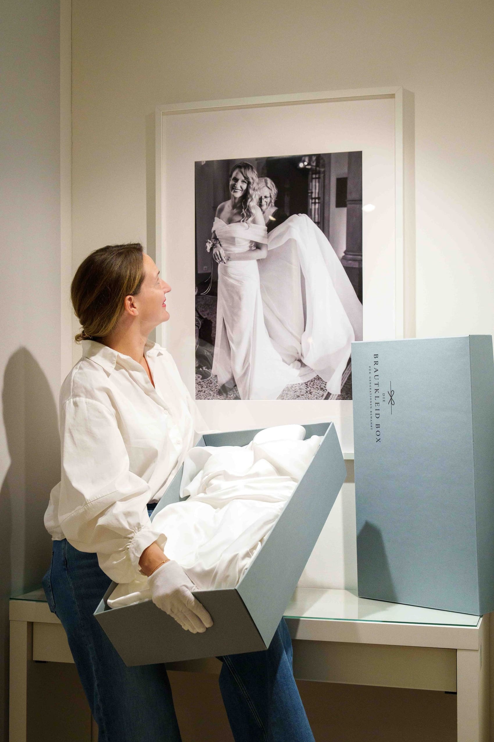 Frau legt ihr Brautkleid in die Brautkleidbox, im Hintergrund ein Hochzeitsfoto von Braut und Mutter
